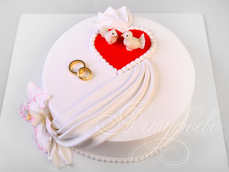 Белый свадебный торт с красным сердечком, голубями, свадебными кольцами и драпировкой